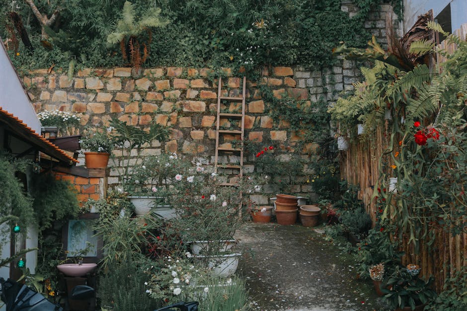 A narrow garden space situated in a tight terrace, featuring a gravel pathway bordered by diverse plants and garden pots. On the left, there are various potted plants placed on shelves and the ground,